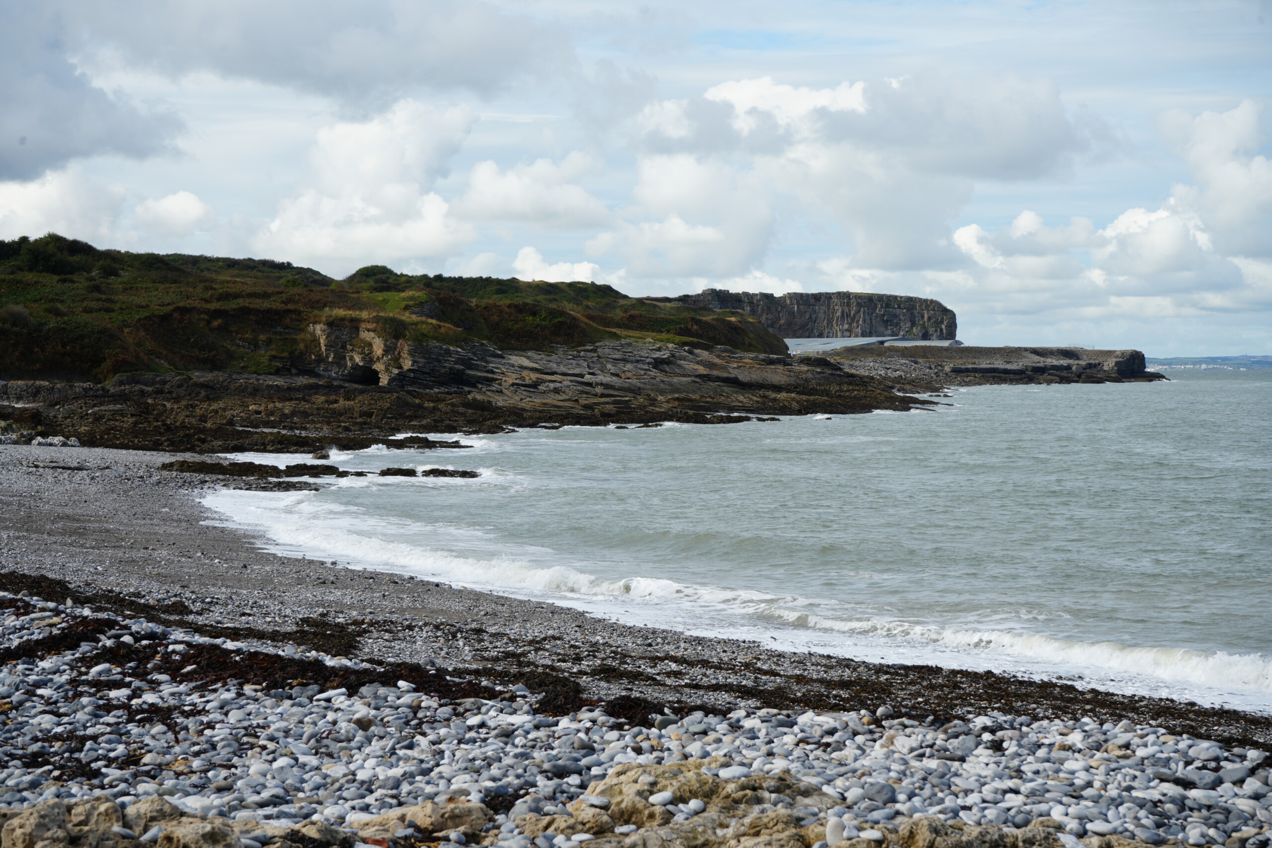Beach_Penmon_Point Rocky pebble beach at Penmon Point in Anglesey, with rugged cliffs and greenery in the background, and gentle waves breaking along the shore under a partly cloudy sky.