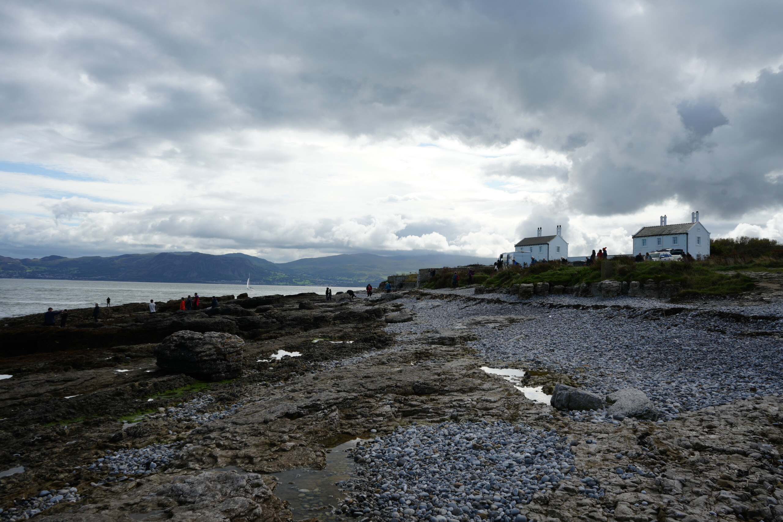 Edward_Hopper_Houses_Penmon_Point Rocky shoreline at Penmon Point with scattered visitors, two white cottages in the background, and mountains across the water under a cloudy sky.