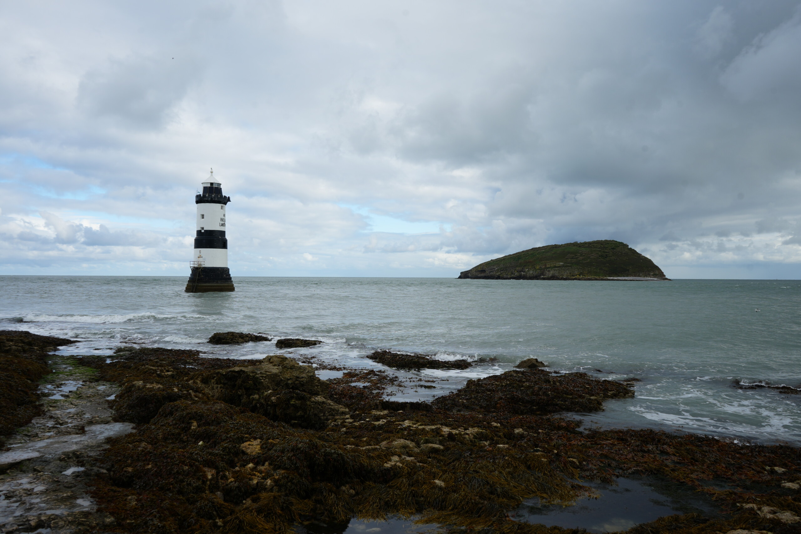 Penmon_Point_And_Puffin_Island Penmon Point Lighthouse standing in the sea near the rocky shoreline, with Puffin Island visible in the background under a cloudy sky.
