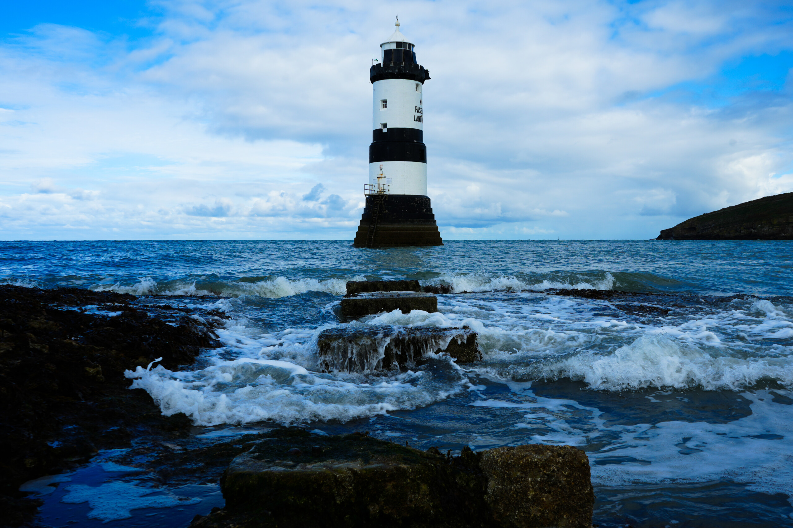 Penmon_Point_Lighthouse_North_Wales Penmon Point Lighthouse in North Wales, surrounded by crashing waves against rocky steps in the foreground, under a partly cloudy blue sky.