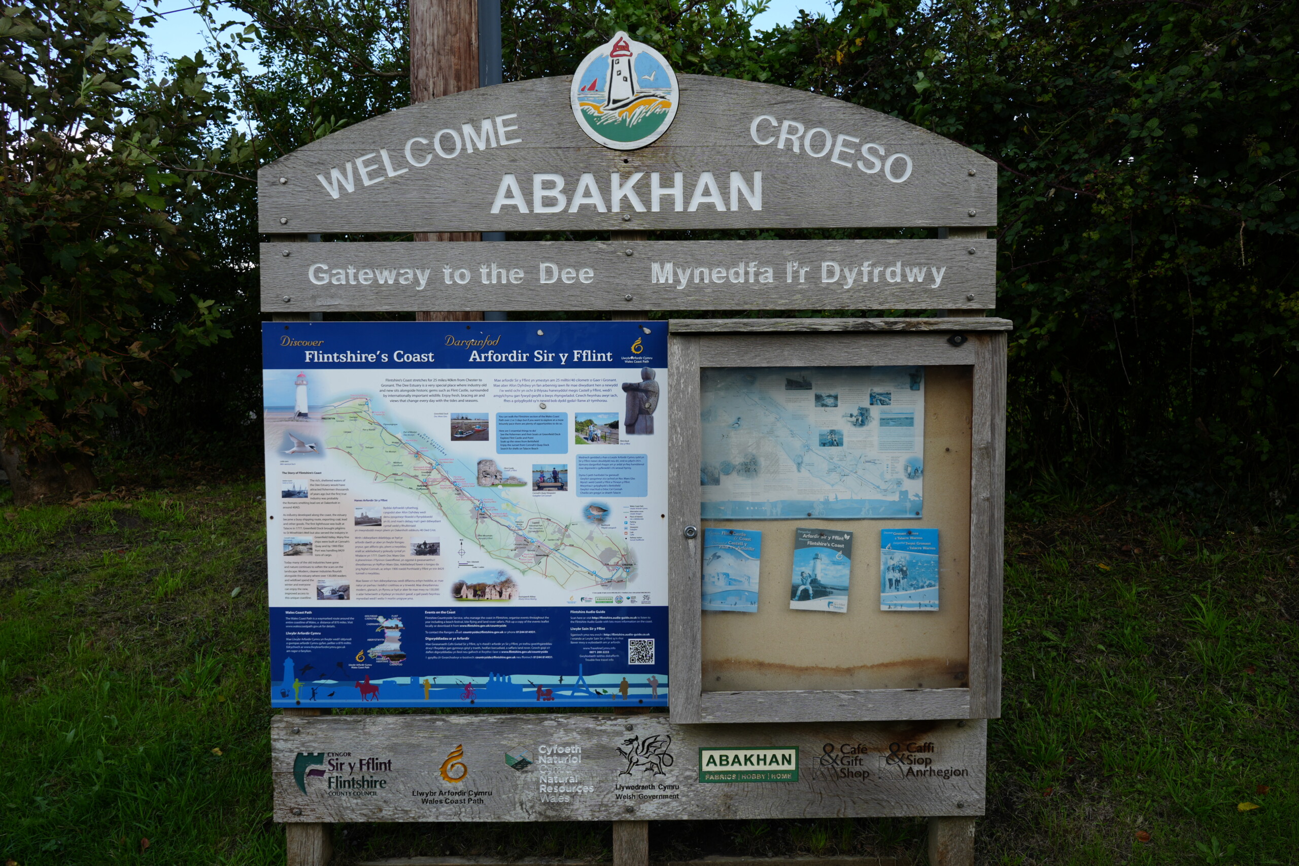 Wooden welcome sign in the car park near the Duke of Lancaster ship in Flintshire. The sign reads 'Welcome to Abakhan – Gateway to the Dee' and includes a map of Flintshire’s Coast, local information, and tourist leaflets, surrounded by trees and greenery.