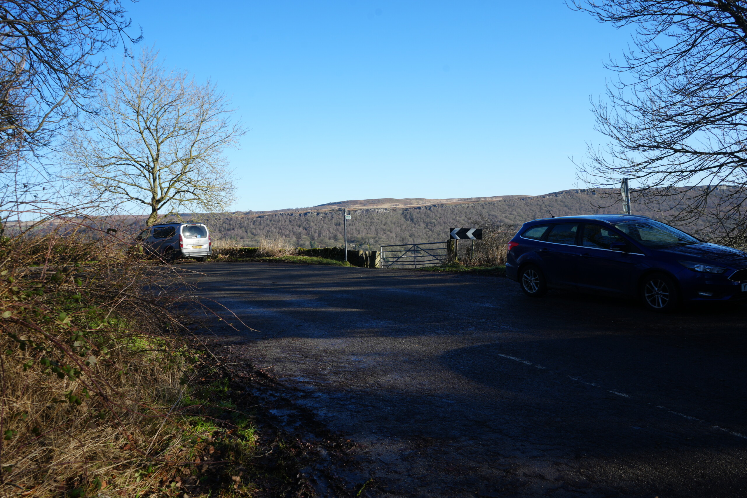 Cross_Road_TOwards_Path A quiet rural road junction in Eyam, England, with two parked cars—one silver and one blue—overlooking rolling hills and woodland in the distance. A large, leafless tree stands against a bright blue sky, casting long shadows on the tarmac. A metal gate and road sign with black and white chevrons indicate a sharp turn ahead. The scene captures the peaceful countryside with open views across the valley.