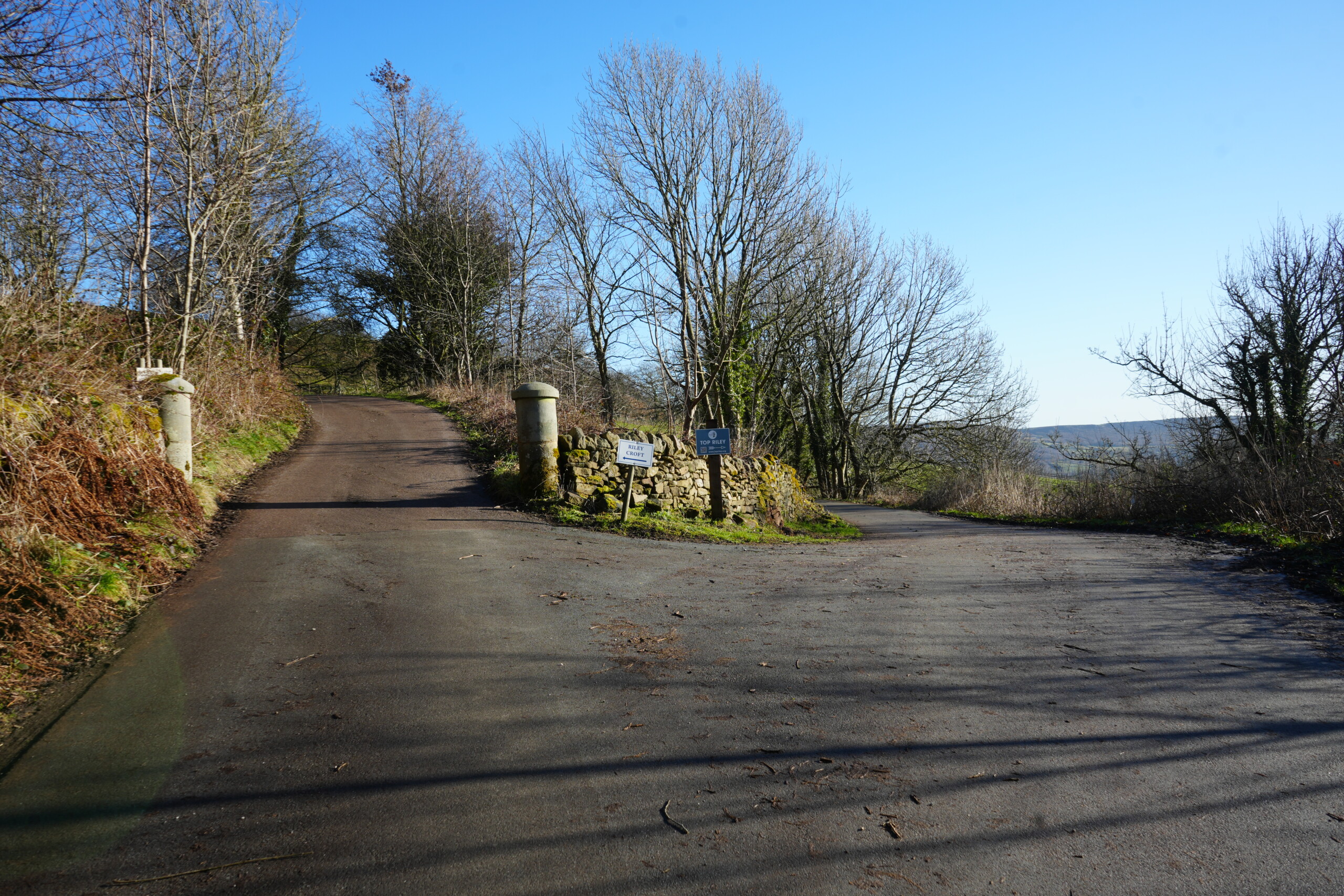 Fork_Right_To_Riley_Graves A rural road junction in Eyam, England, where the path forks in two directions. The left road ascends a hill between two stone pillars, leading towards a private estate, while the right road curves gently downhill towards the historic Riley Graves. A dry-stone wall with moss-covered stones separates the two paths, featuring signposts indicating directions. The surrounding landscape is dotted with leafless trees, undergrowth, and distant rolling hills beneath a bright blue sky.