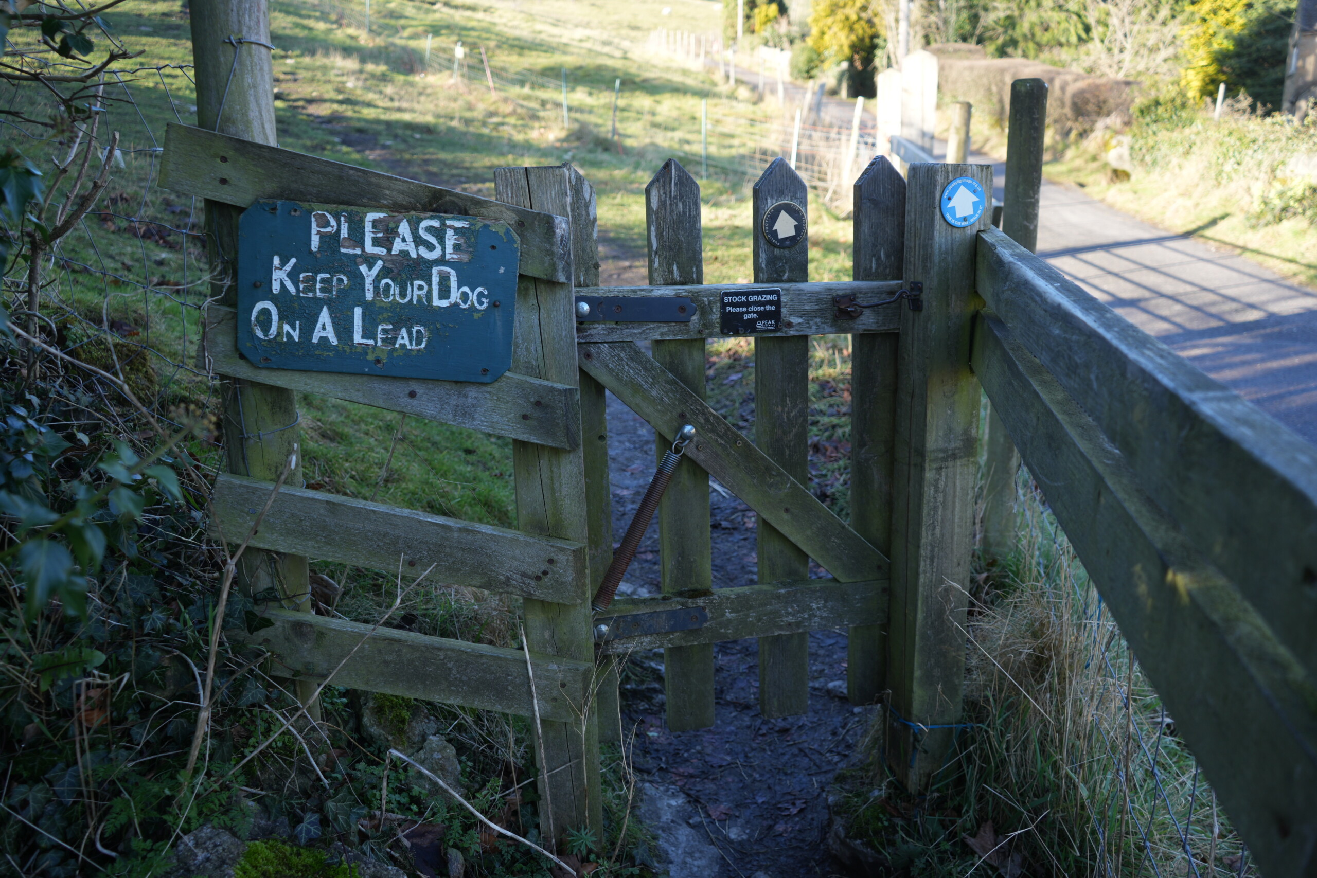 Go_Through_Gate A wooden gate leading to a countryside footpath, with a green sign reading 'PLEASE KEEP YOUR DOG ON A LEAD' attached to the fence. The gate is secured with a metal latch and a spring mechanism, and there are two waymarker signs with house-shaped symbols indicating a walking route. A small black sign on the gate warns of stock grazing and reminds walkers to close the gate behind them. Beyond the gate, a dirt path leads uphill through open fields, enclosed by fencing. Sunlight casts shadows across the nearby tarmac road.