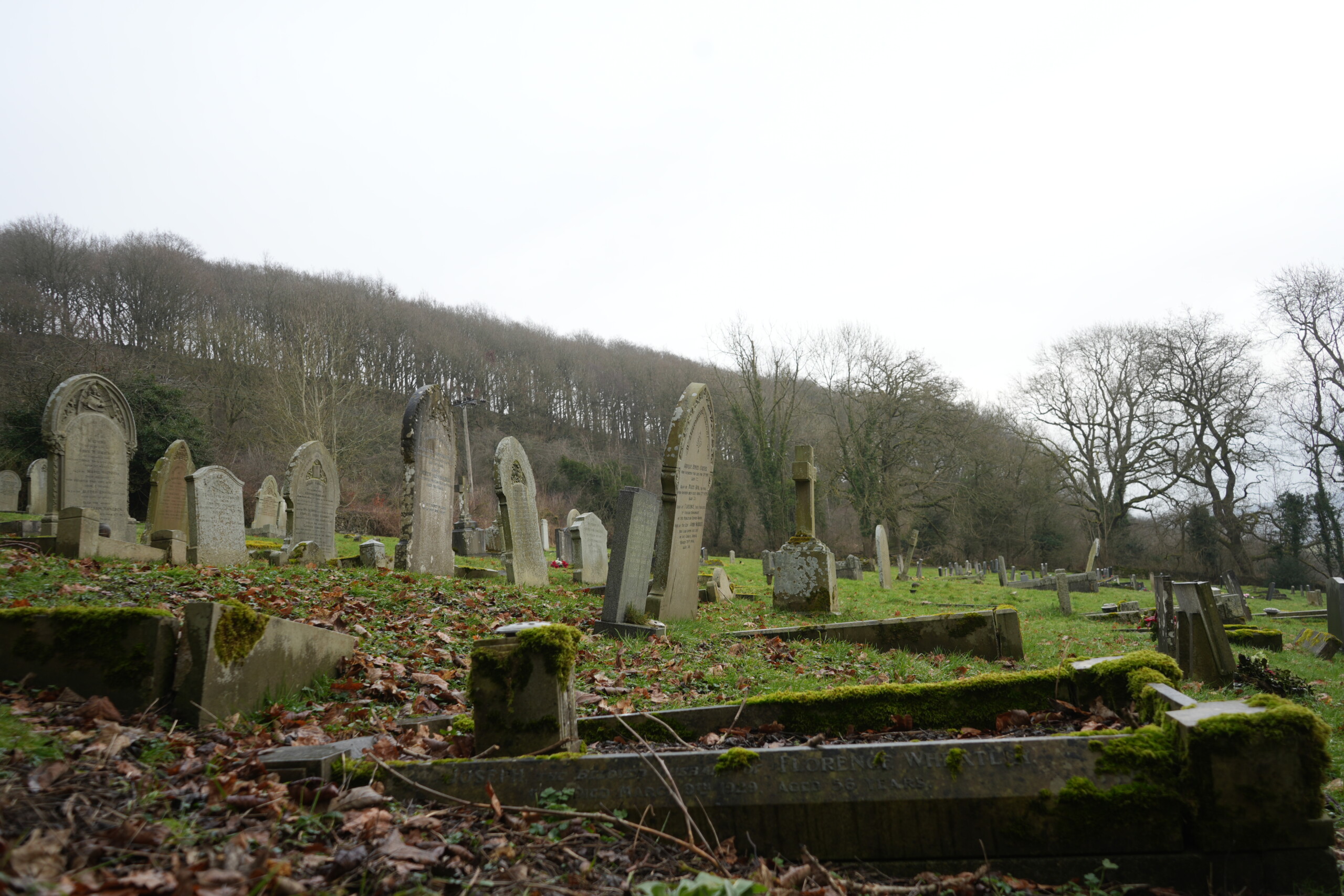 Grave_Yard_Along_Path A historic graveyard in Stoney Middleton, England, with weathered and moss-covered headstones leaning at various angles. The ground is covered in fallen leaves and patches of grass, creating a peaceful yet solemn atmosphere. Some gravestones feature intricate carvings, while others are worn and eroded by time. Leafless trees and a wooded hillside in the background add to the sense of history and remembrance in this resting place, which is closely linked to the village’s tragic plague history.