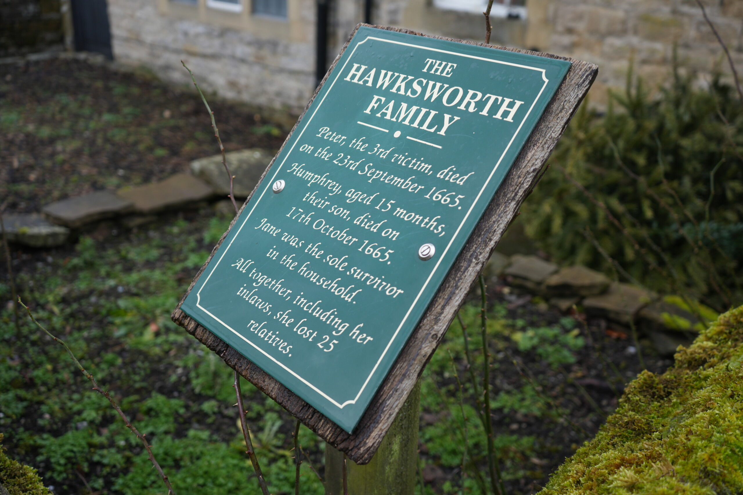 Hawksworth_Family_Eyam A weathered green informational sign titled 'The Hawksworth Family' stands in front of a historic stone cottage in Eyam, England. The sign details the tragic loss of Peter Hawksworth, who died in September 1665, and his 15-month-old son, Humphrey, who passed away in October 1665, during the village's plague outbreak. Jane Hawksworth was the only household survivor, having lost 25 relatives in total. The sign is mounted on a wooden post, surrounded by moss-covered stone walls and a garden with sparse greenery.