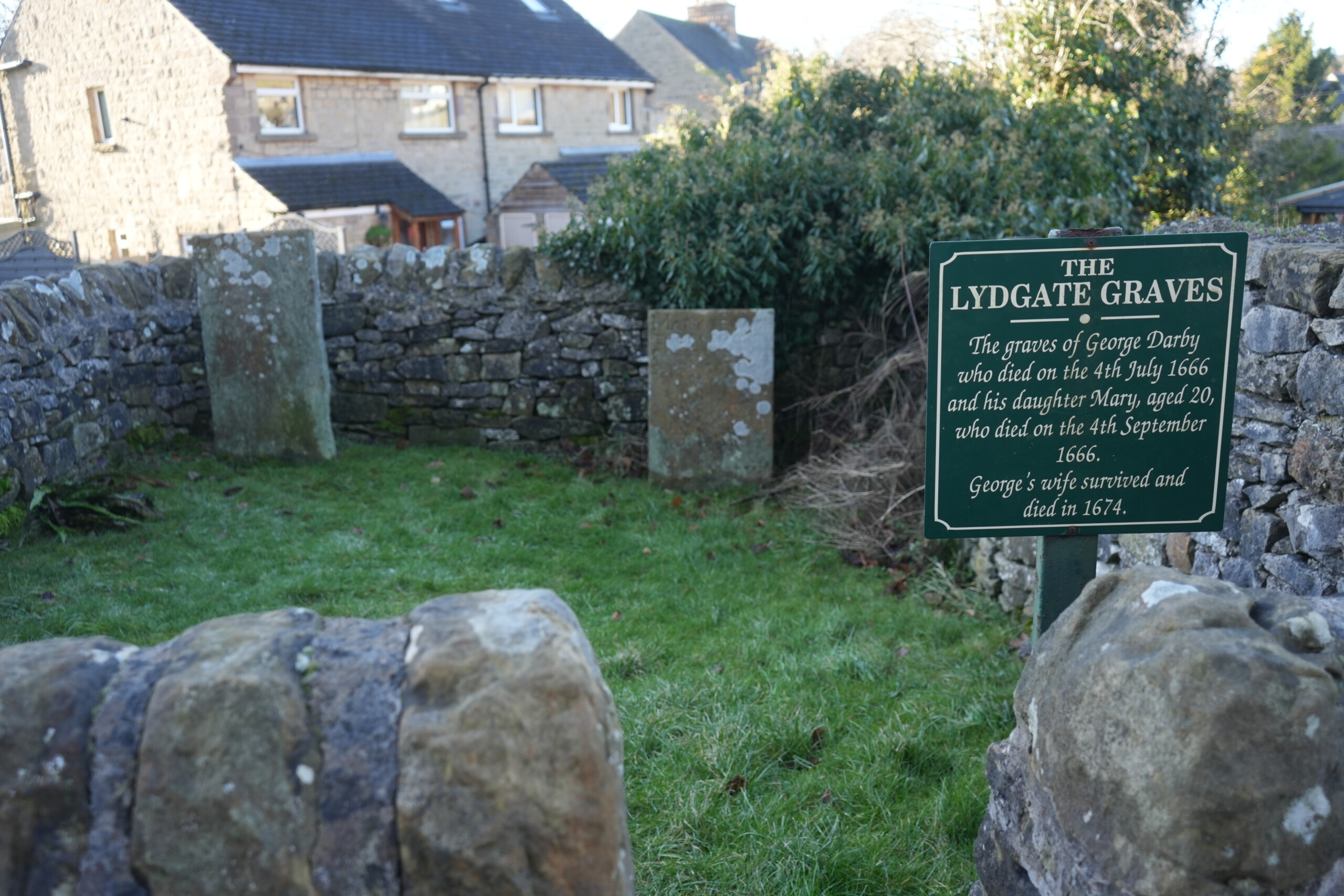Lydgate_Graves_Eyam A small, enclosed grave site with two weathered gravestones standing within a stone-walled area, surrounded by grass. A green sign with white lettering titled 'The Lydgate Graves' provides historical information about George Darby and his daughter Mary, who both died in 1666. Modern houses can be seen in the background beyond the stone wall.