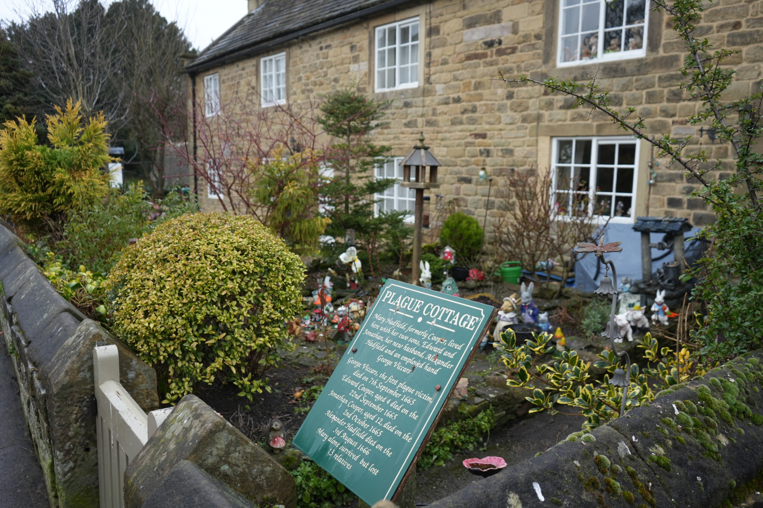 Plague_Cottage_Eyam A historic stone cottage in Eyam, England, known as 'Plague Cottage,' with a green informational sign in the foreground detailing its connection to the 1665 plague. The cottage features white-framed windows and a small front garden filled with decorative ornaments, shrubs, and a birdhouse. The stone wall surrounding the property is covered in moss, with a cream-coloured wooden gate at the entrance.