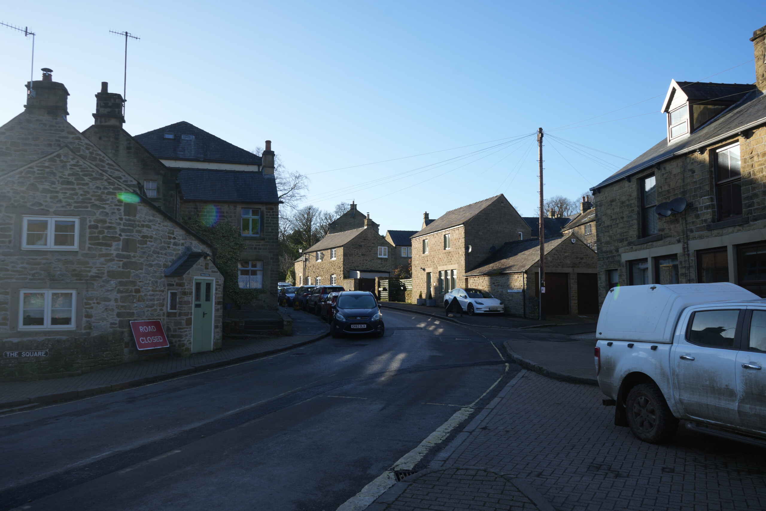 Retrace_Steps_Back_To_The_Car A quiet village street lined with traditional stone houses in warm sunlight. Parked cars are seen along the road, and a red 'Road Closed' sign is positioned near a house with a pale green door. Overhead wires stretch across the scene, with clear blue skies above. The setting appears to be a historic English village.