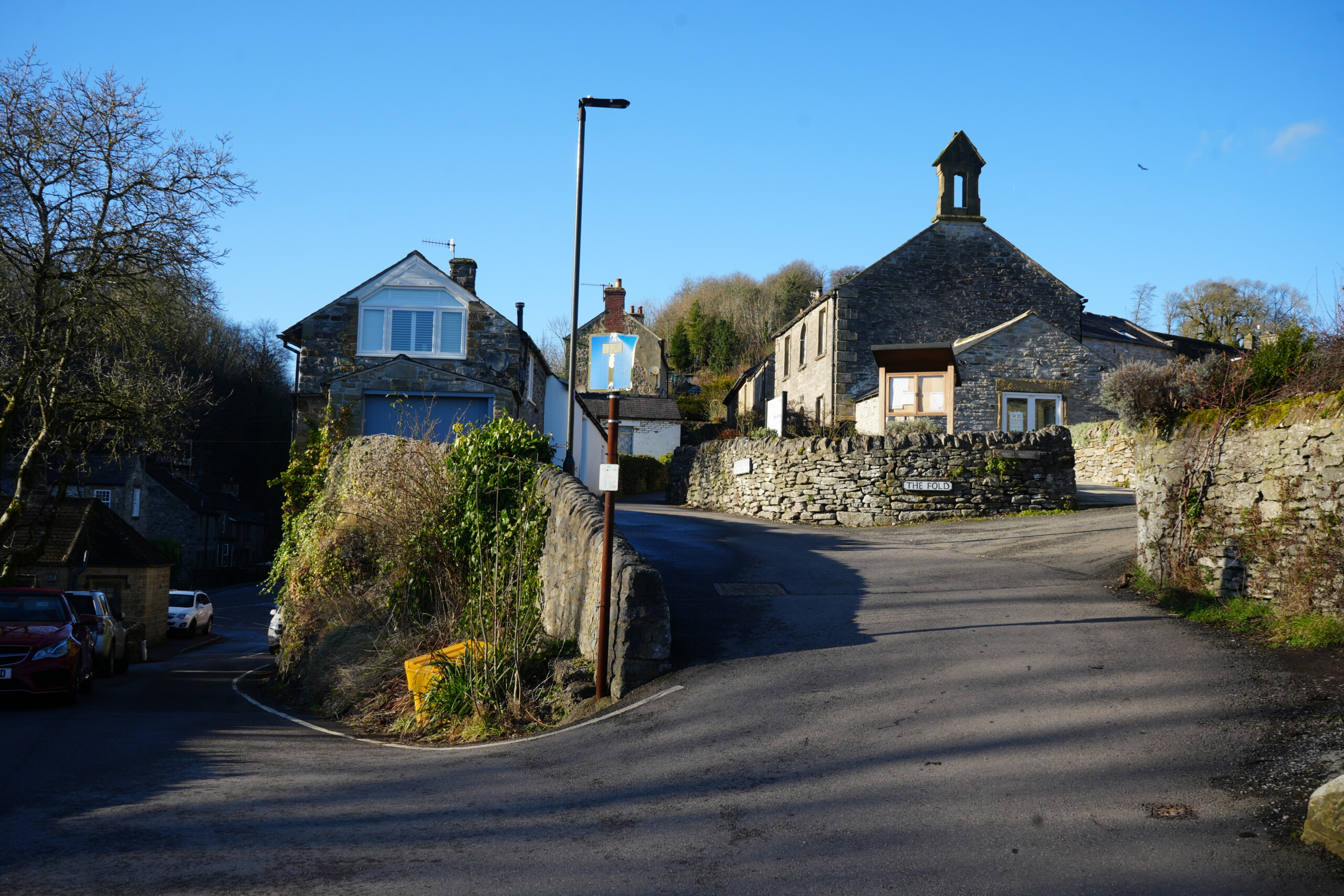 Right_Hand_Fork A picturesque fork in the road in a historic English village, featuring traditional stone cottages and dry-stone walls. The right-hand path leads uphill towards 'The Fold,' a stone building with a small bell tower, while the left path curves gently past ivy-covered walls and a modern house with white shutters. A lamppost with a faded blue sign stands at the junction, and a yellow grit bin is positioned near the stone wall. The clear blue sky and long shadows suggest a bright winter's day in a quiet countryside setting.