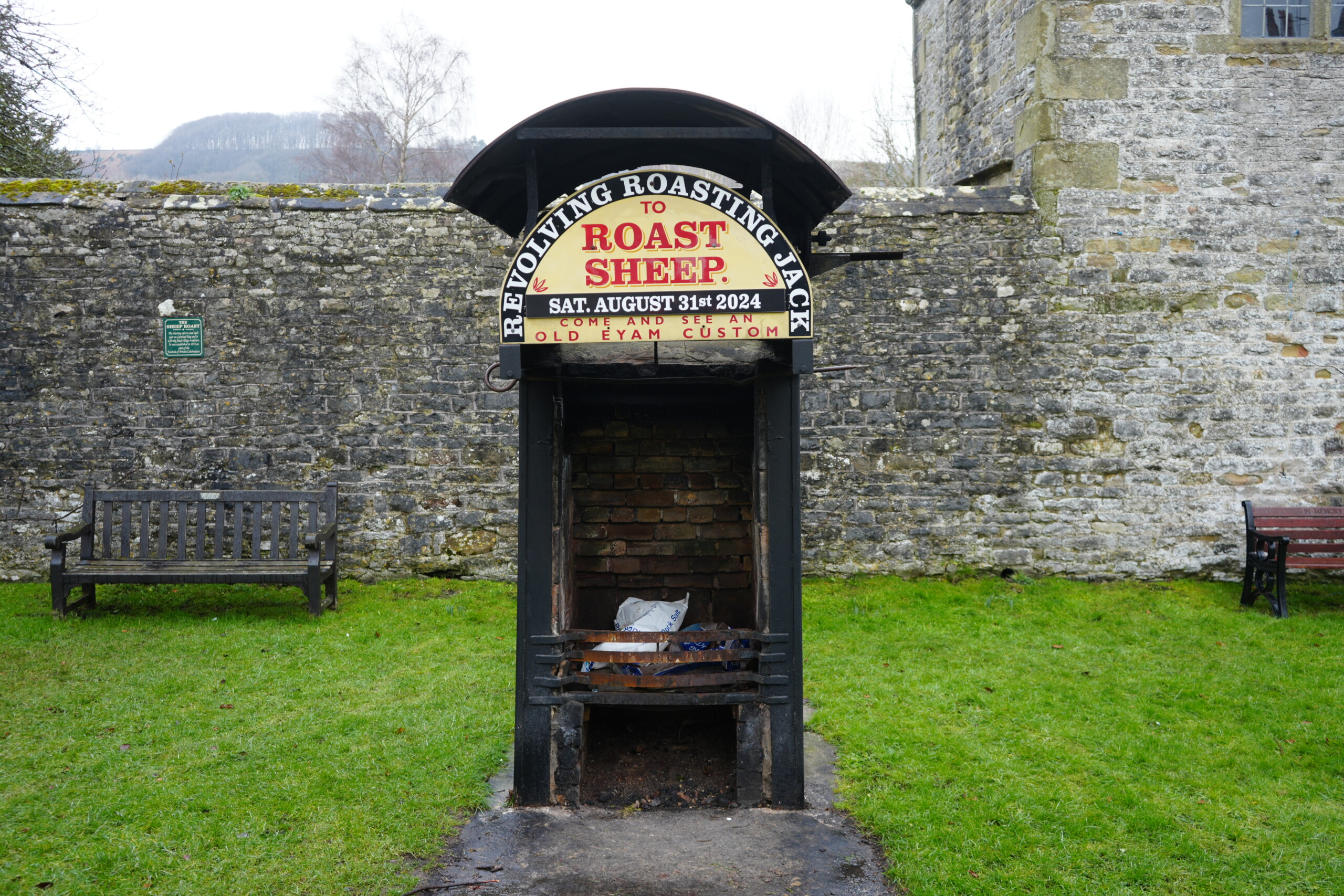 Roast_Sheep_Eyam A historic revolving roasting jack in Eyam, England, featuring a black metal structure with a brick interior, traditionally used for roasting sheep. A sign above reads 'Revolving Roasting Jack to Roast Sheep' with a date for an event on August 31st, 2024, inviting visitors to witness an old Eyam custom. The roasting jack is set against a stone wall, with wooden benches on either side and a grassy area in the foreground. A distant hill and trees are visible in the background.