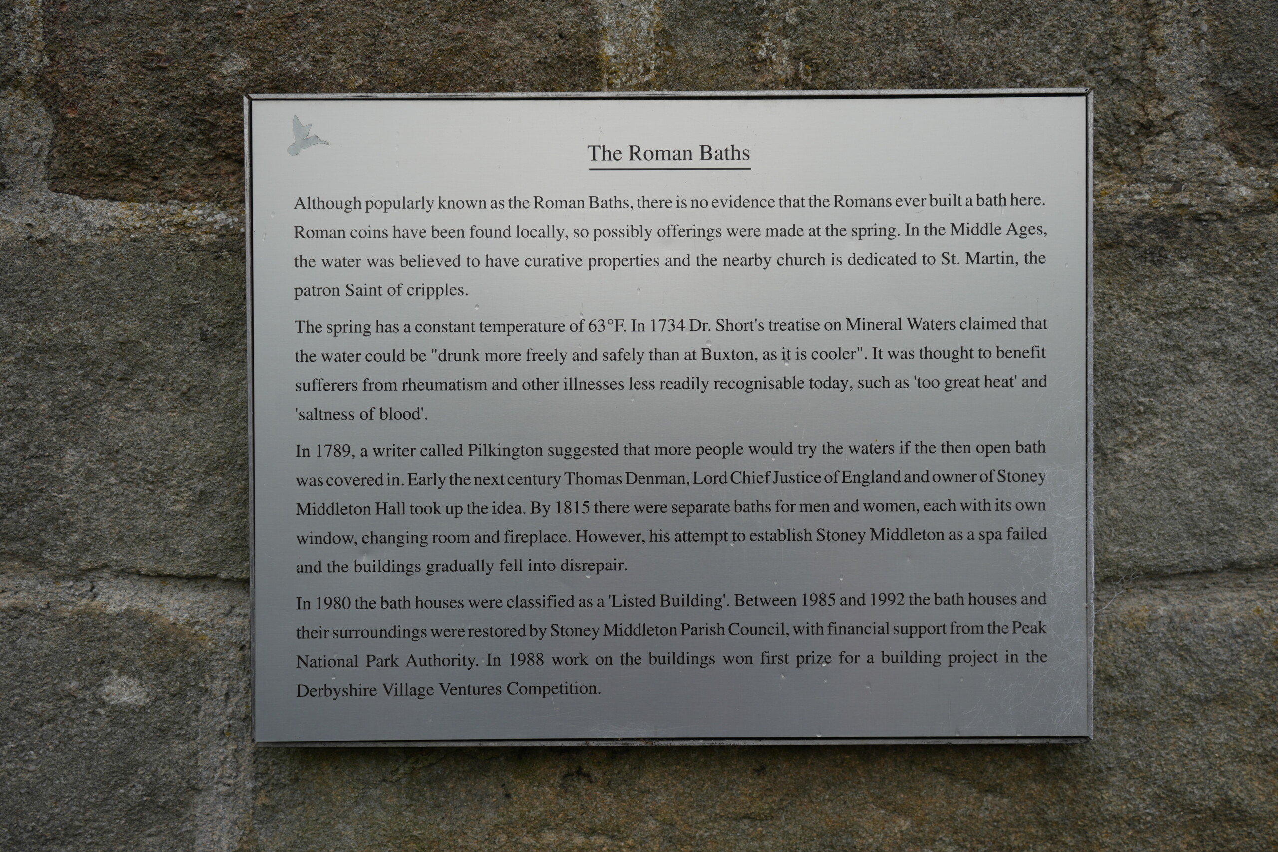 Roman_Baths_Stoney_Middleton A historical information plaque mounted on a stone wall, detailing the history of the Roman Baths in Stoney Middleton, England. The sign explains that, despite the name, there is no evidence of Roman-built baths at the site, though Roman coins were found nearby. It describes the medieval belief in the spring’s healing properties and mentions its consistent temperature of 63°F. The plaque also recounts attempts in the 18th and 19th centuries to establish a spa, which ultimately fell into disrepair, before the baths were restored and classified as a listed building in 1980.