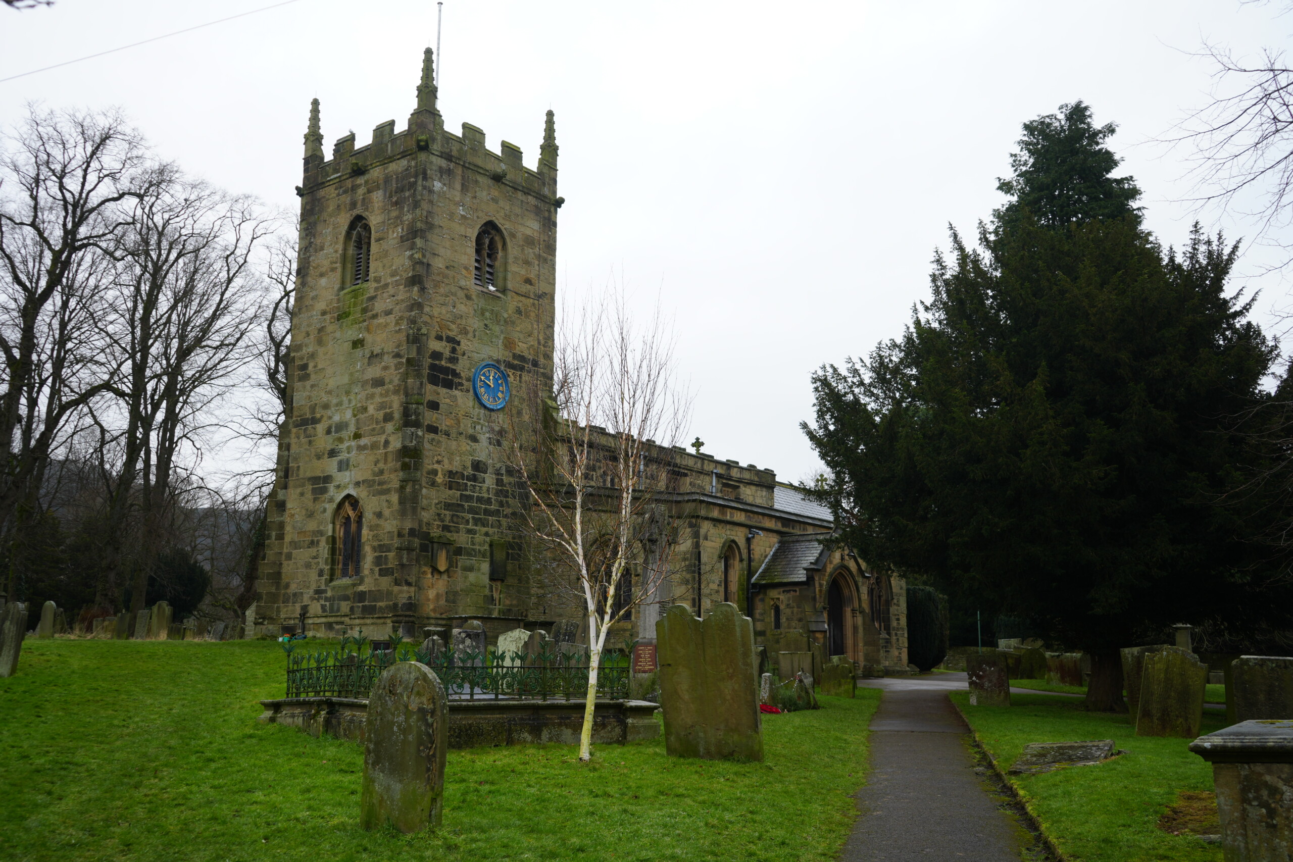 St_Lawrence_Church_Eyam St. Lawrence Church in Eyam, England, a historic stone church with a square tower featuring battlements and a blue clock face. The churchyard is filled with weathered gravestones, including a prominent white-barked tree near the entrance. A pathway leads towards the church's arched doorway, surrounded by lush green grass and mature trees. The overcast sky adds a sombre atmosphere to the scene, reflecting the church’s historical significance in the village's past, particularly during the plague era.