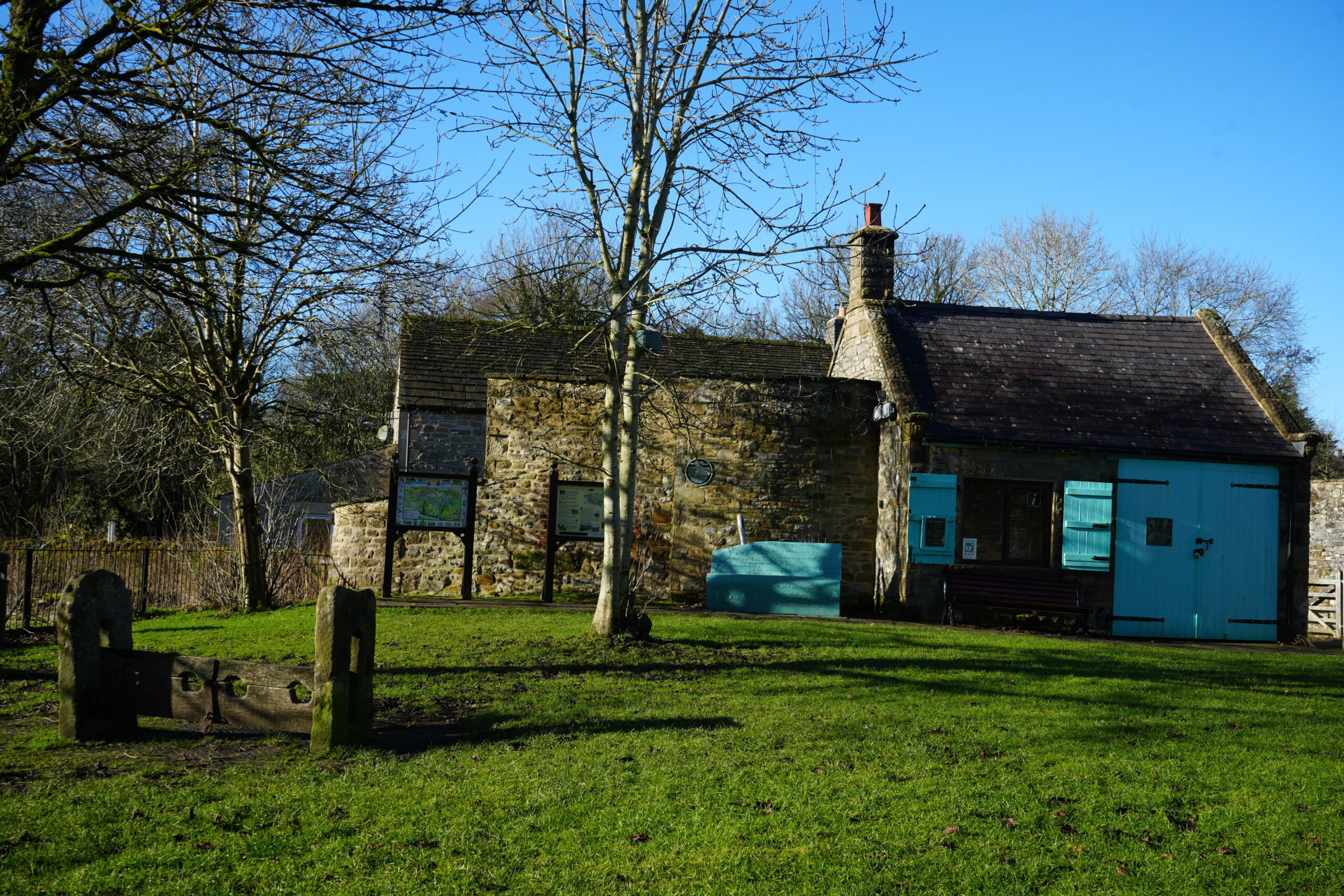Stocks_Eyam_And_Visitor_Centre A historic stone building in Eyam, England, with blue wooden shutters and doors, situated in a grassy area with old wooden stocks in the foreground. The site serves as a visitor centre, with informational boards mounted on stands near the entrance. Leafless trees frame the scene, and the bright blue sky contrasts with the rustic stone architecture. The stocks, a remnant of historical punishment methods, add to the historical significance of the location.