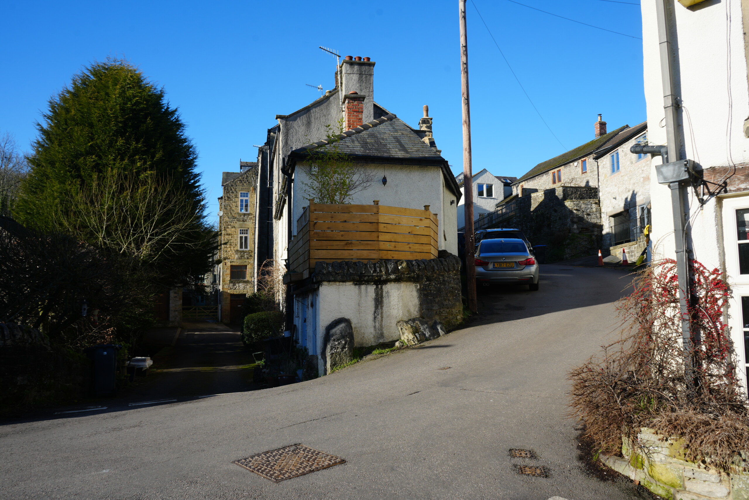 T-Junction_To_Eyam A scenic village junction in Stoney Middleton, England, featuring a narrow road splitting in two directions. A historic whitewashed cottage with a stone base and a wooden balcony is perched on the corner, elevated on a rocky foundation. To the right, a steep road leads uphill past more traditional stone buildings, with a parked car visible. On the left, a shaded driveway leads to additional village houses. The scene is framed by a tall evergreen tree and a blue sky, with warm sunlight casting shadows on the buildings and road.