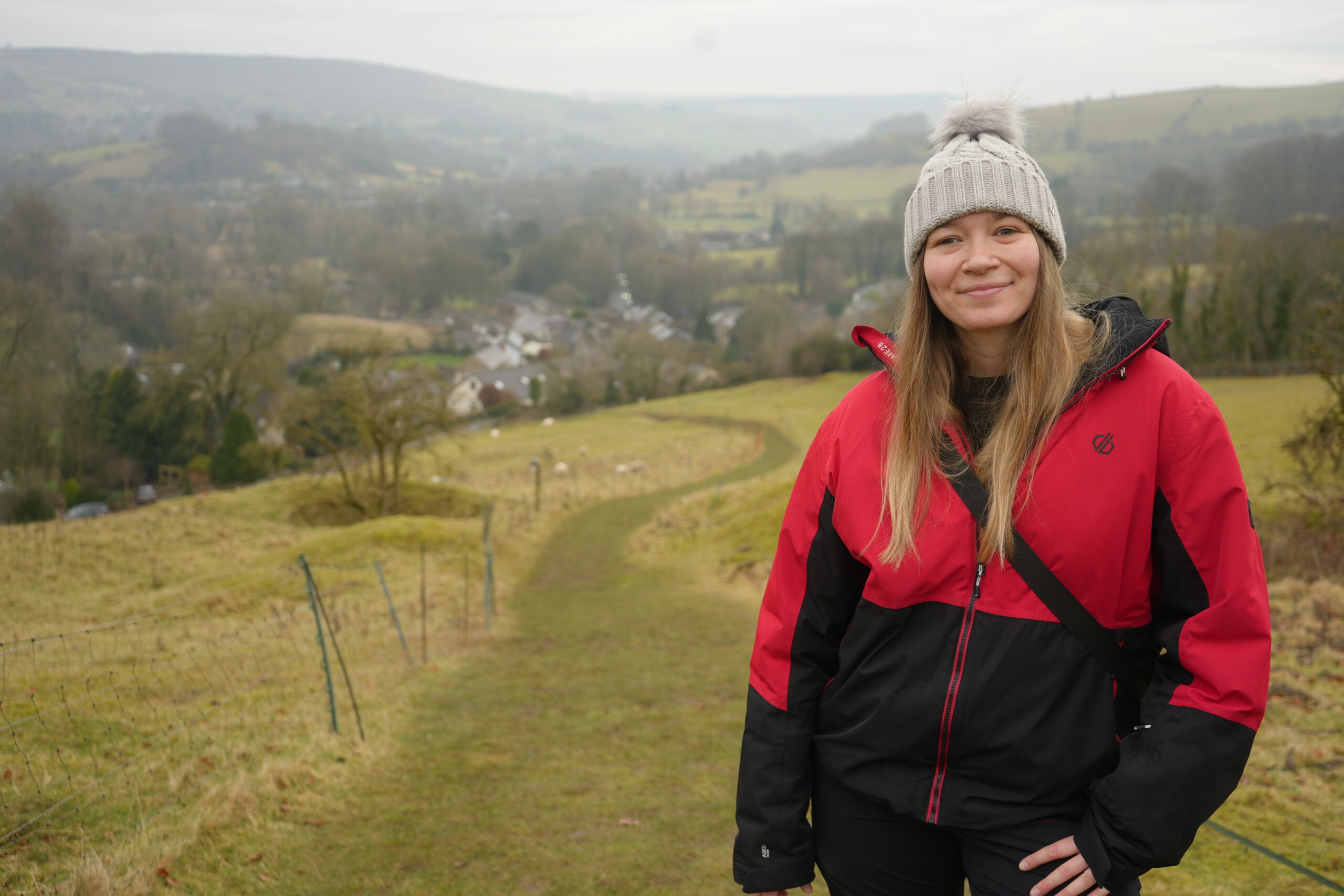 Take_In_The_Views The Nott'm Lass wearing a red and black outdoor jacket, black trousers, and a grey knitted beanie with a fluffy pom-pom stands smiling on a grassy hillside. Behind her, a winding footpath leads down towards a scenic countryside village, with rolling hills, scattered trees, and sheep grazing in the fields. The sky is overcast, creating a soft, diffused light over the landscape.