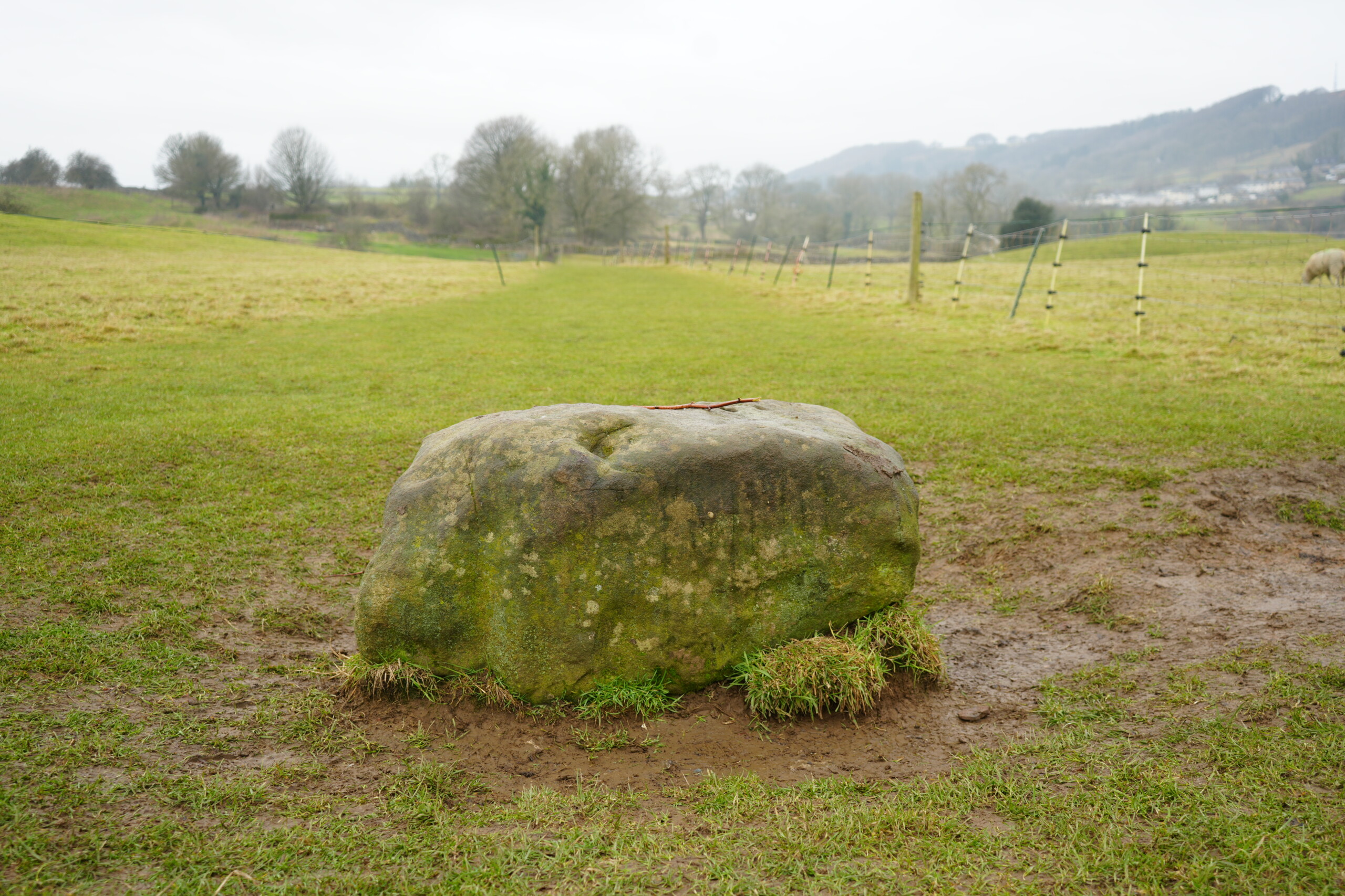 The_Boundary_Stone A moss-covered stone known as the Boundary Stone sits prominently in a grassy field, surrounded by muddy ground. A pathway extends into the distance, flanked by wooden fence posts and wire fencing. Rolling hills, scattered trees, and a misty countryside landscape can be seen in the background under an overcast sky.
