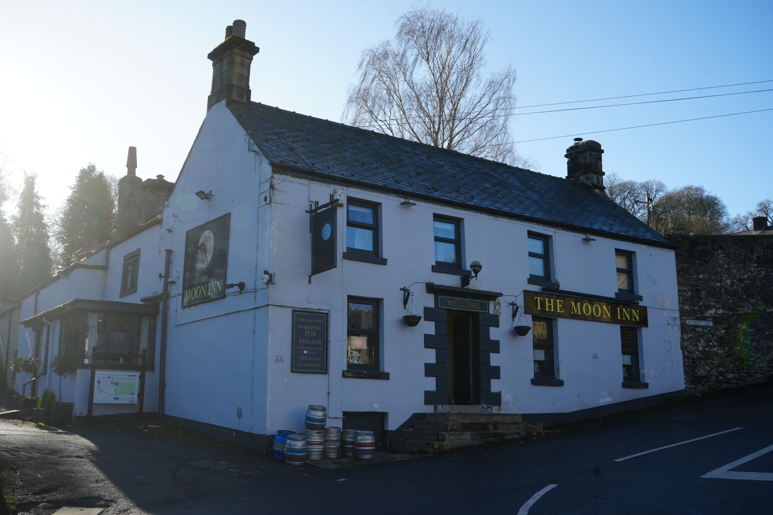 The_Moon_Inn_Stoney_Middleton The Moon Inn, a traditional pub in Stoney Middleton, England, housed in a historic white-painted building with dark grey window frames and signage. The pub's name is displayed in gold lettering above the entrance and on a hanging sign featuring an image of the moon. A row of beer barrels sits outside near the entrance, and a small porch with an informational board is visible on the left side. The pub is situated at a road junction, with a stone wall and leafless trees in the background, under a clear blue sky.