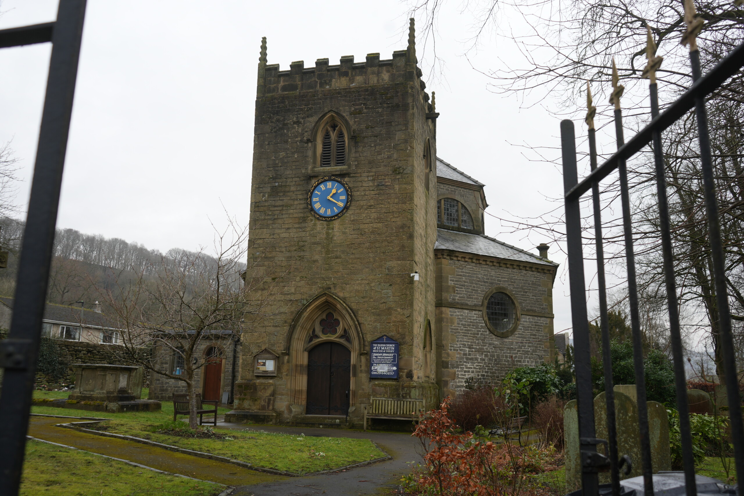 The_Parish_Church_Of_St_Martin_Stoney_Middleton The Parish Church of St. Martin in Stoney Middleton, England, a historic stone church with a square tower featuring battlements and a blue clock face with gold Roman numerals. The entrance has a pointed arch doorway, and a church sign is mounted on the wall beside it. The churchyard is surrounded by a black wrought iron fence, with gravestones, a stone tomb, and neatly maintained gardens. Leafless trees and a hillside covered in woodland create a picturesque backdrop on this overcast day.