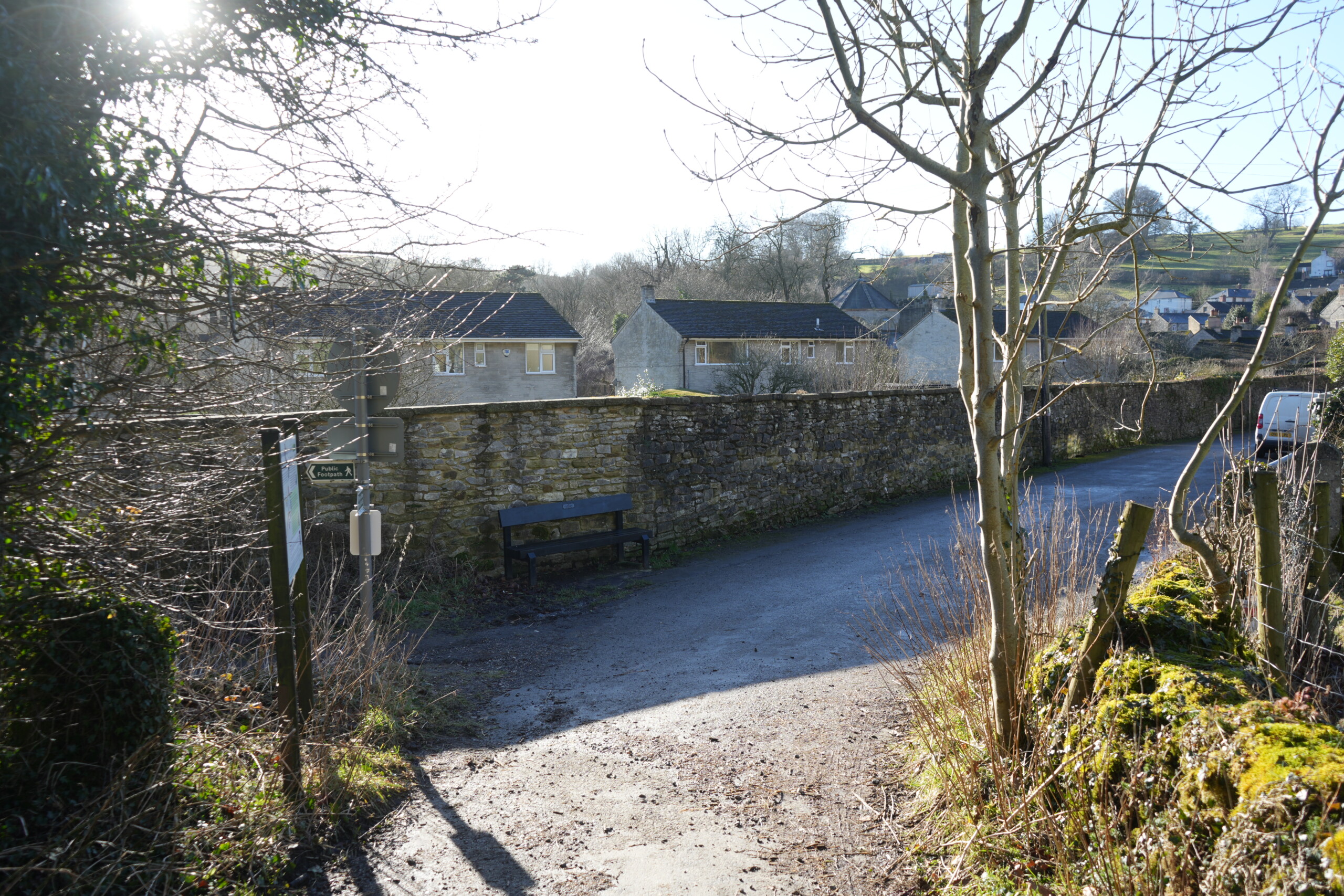 Turn_Right_Bottom_Of_Path A quiet village lane in Stoney Middleton, England, where a footpath meets a paved road lined by an old dry-stone wall. A wooden bench is placed against the wall, providing a resting spot for walkers. Sunlight filters through the bare branches of nearby trees, casting long shadows on the ground. In the background, stone cottages and rolling countryside stretch towards the horizon, creating a peaceful rural scene. A white van is partially visible on the right, parked along the roadside." Let me know if you need any modifications!