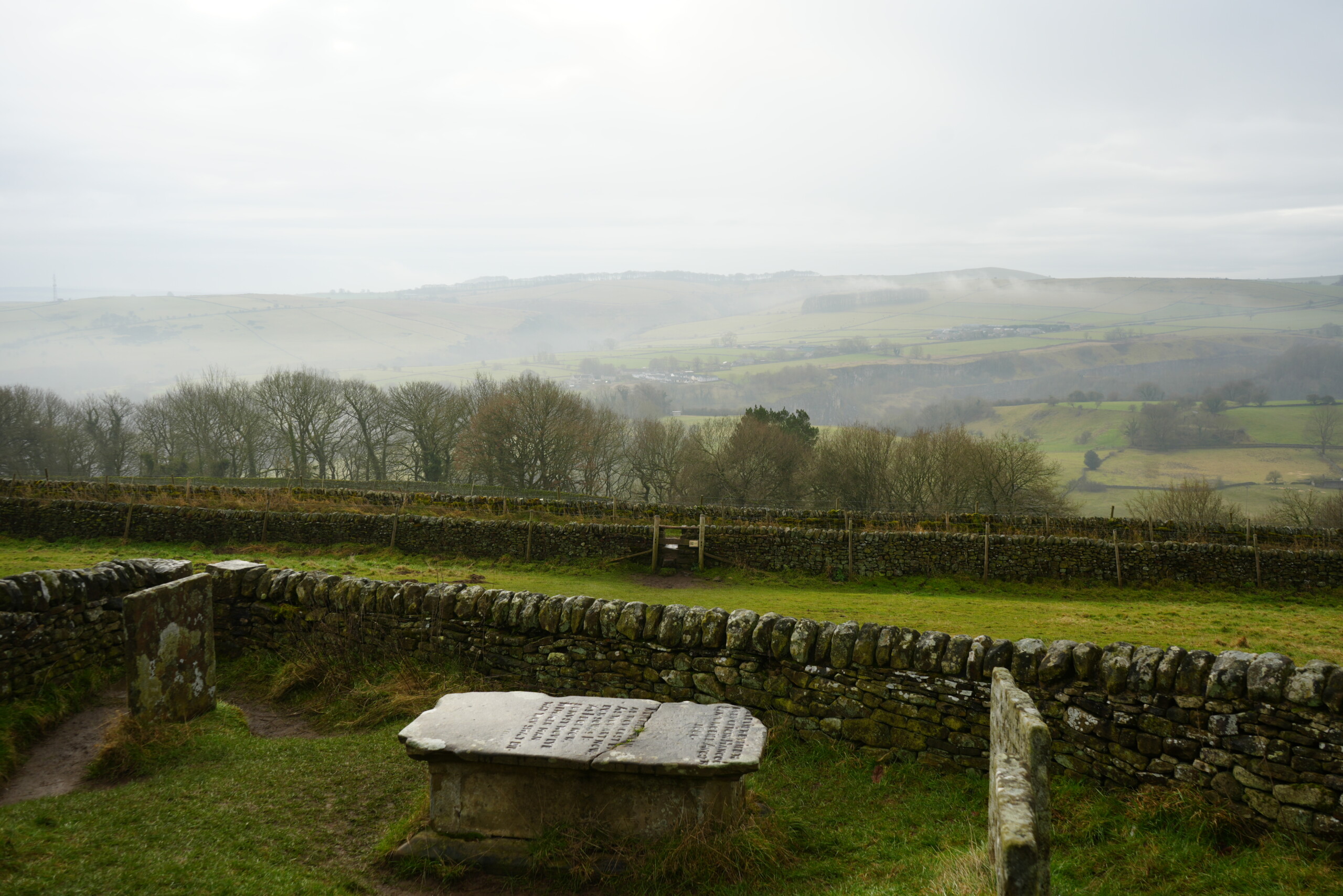 View_From_Riley_Graves A misty, panoramic view from the Riley Graves in Eyam, England, overlooking rolling green hills and patchwork fields in the distance. In the foreground, a weathered stone table tomb and gravestones sit within a low dry-stone wall enclosure. Beyond the graves, additional stone walls divide the landscape, and a wooden stile provides access to the surrounding countryside. The scene is framed by leafless trees, with mist clinging to the distant hills, creating a peaceful yet poignant atmosphere.
